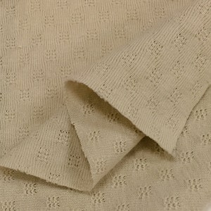 May include: Close-up of a beige knitted fabric with a textured pattern. The fabric is folded in half, showing the texture of the knit.
