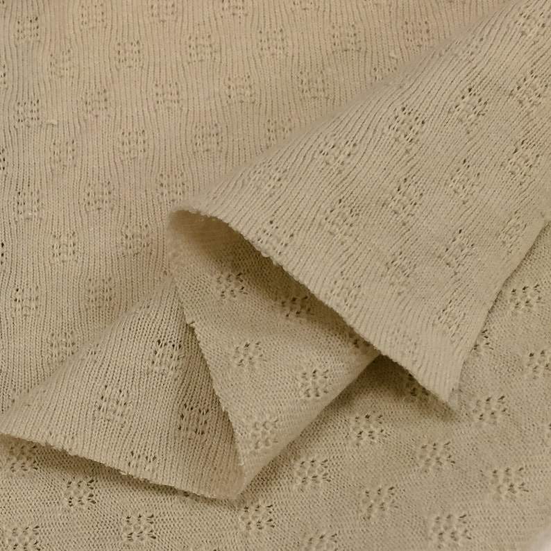 May include: Close-up of a beige knitted fabric with a textured pattern. The fabric is folded in half, showing the texture of the knit.