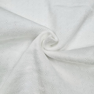 May include: Close-up of white fabric with a textured, open-weave design. The fabric is tightly woven with small, evenly spaced holes, creating a delicate, airy appearance. The material is soft and appears to be folded, showing the texture and drape of the fabric.