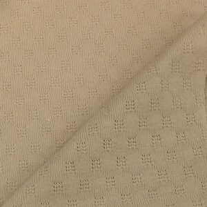 May include: Close-up of a beige knitted fabric with a small, repeating pattern of holes.