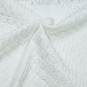 May include: Close-up of white fabric with a textured, woven pattern. The fabric is draped, showing folds and shadows. The weave creates a grid-like design with small, repeating details.