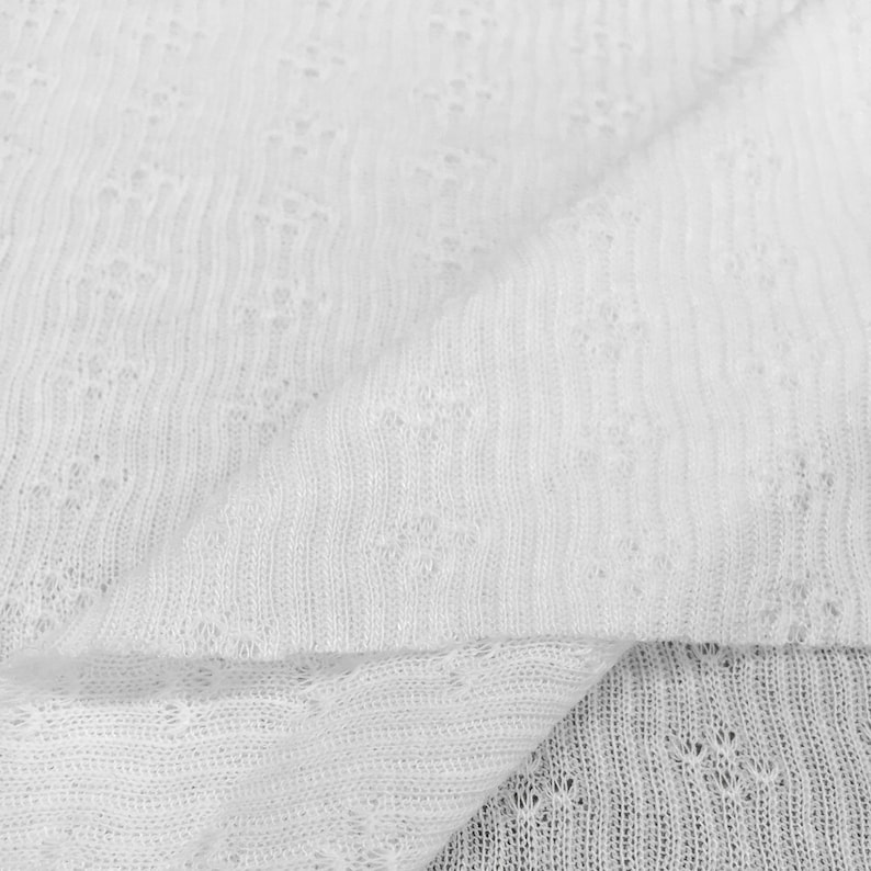 May include: Close-up of white fabric with a delicate, open weave. The material is folded, showing the texture and pattern of the knit. The fabric appears soft and lightweight, suitable for clothing or accessories.