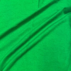 May include: Bright green fabric with visible folds and a soft texture. The fabric's vibrant color and subtle shadows create a sense of depth and movement. The material appears to be lightweight and flowing, suitable for clothing or decorative purposes.