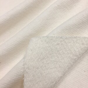 May include: Close-up of a white fabric with a soft, fuzzy texture on one side and a smooth, woven texture on the other side.