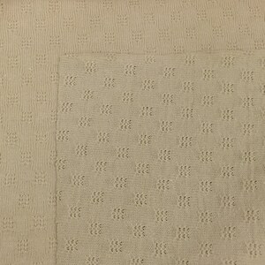 May include: A close-up of a beige knitted fabric with a textured pattern. The fabric has a subtle sheen and a soft, comfortable feel.