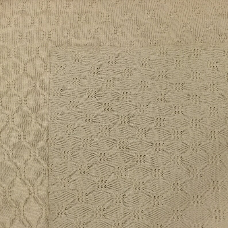 May include: A close-up of a beige knitted fabric with a textured pattern. The fabric has a subtle sheen and a soft, comfortable feel.