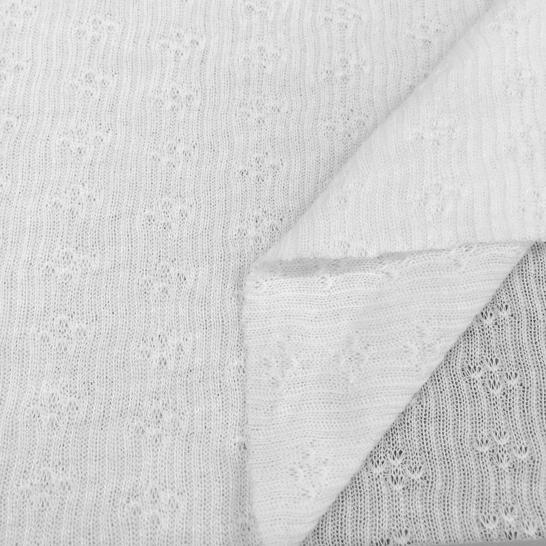 May include: White knitted fabric with a delicate openwork pattern. The fabric is folded in half, showing the texture and detail of the knit.