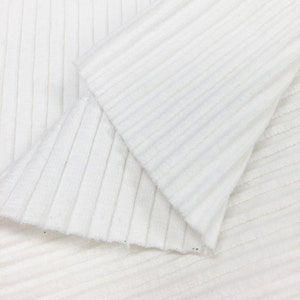 Op de afbeelding: Witte ribbelstof met een textuur. De stof is dubbelgevouwen, waardoor de binnen- en buitenkant van het materiaal zichtbaar is.