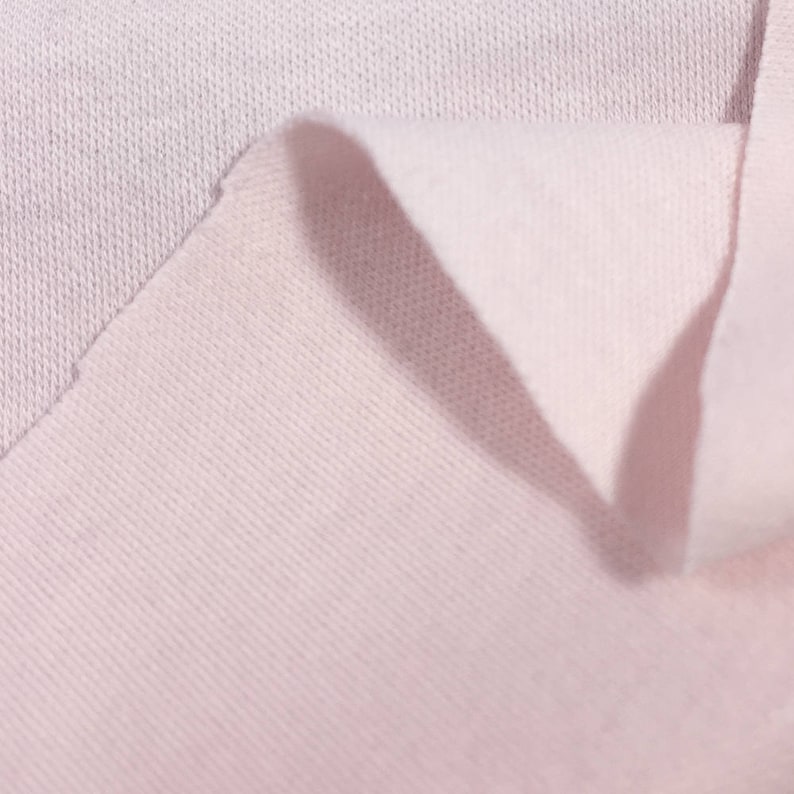 May include: Close-up of a soft, pale pink fabric with a textured surface. The fabric is folded over, revealing the underside.