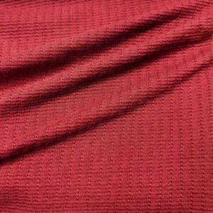 May include: Close-up of a textured, red fabric with a ribbed pattern. The fabric is folded, creating shadows and variations in the color. The material appears soft and possibly knitted or woven.
