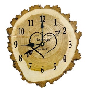 May include: A wooden clock with a heart and arrow design. The clock face is made of a tree slice and has black numbers and hands. The heart has the names "Juan" and "Jenny" and the text "Herrandez est 2014".