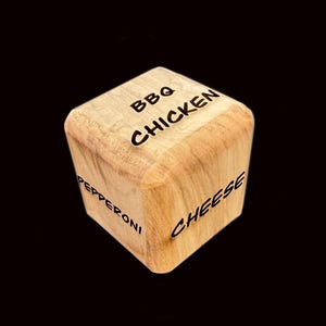 May include: A wooden cube with the words "BBQ CHICKEN", "PEPPERONI", and "CHEESE" etched on its sides. The cube is made of light-colored wood and has rounded edges. The text is in a bold, black font.