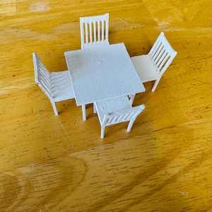 May include: A miniature white dining set, including a square table and four chairs. The chairs have slatted backs, and the table has a textured surface. The set is designed for a dollhouse or similar small-scale setting.