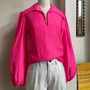 May include: A bright pink long-sleeve blouse with a wide collar and a V-neckline. The blouse has voluminous sleeves and is paired with light-colored denim jeans. The clothing is displayed on a mannequin.
