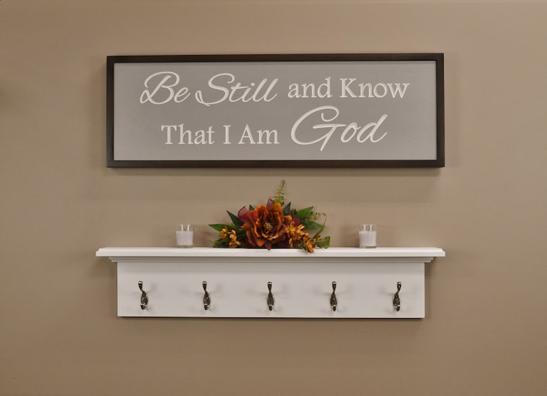 Puede incluir: Estante blanco con cinco ganchos y un letrero enmarcado en marr&oacute;n que dice "Be Still and Know That I Am God". La estanter&iacute;a tiene una moldura decorativa y est&aacute; montada en una pared beige. Un peque&ntilde;o ramo de flores naranja y amarillas se encuentra en la estanter&iacute;a.