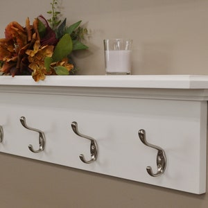 Puede incluir: Estante blanco con cinco ganchos plateados y un borde decorativo. El estante est&aacute; montado en una pared y tiene dos velas en la parte superior.