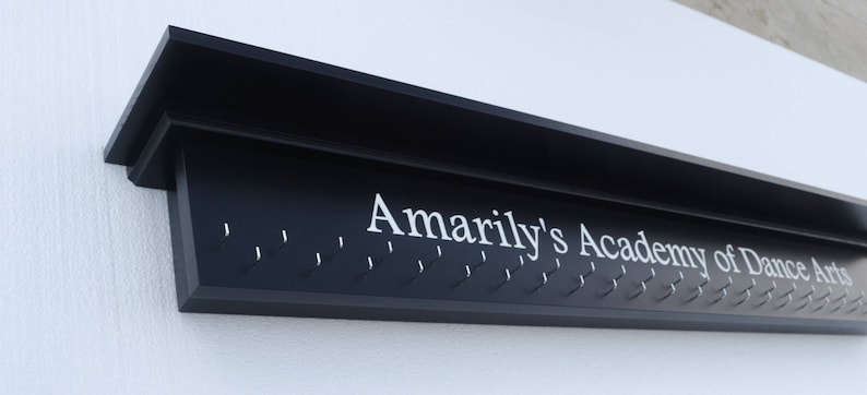 May include: A black wooden wall-mounted medal display with the text "Amarily's Academy of Dance Arts" in white lettering. The display has multiple hooks for hanging medals.