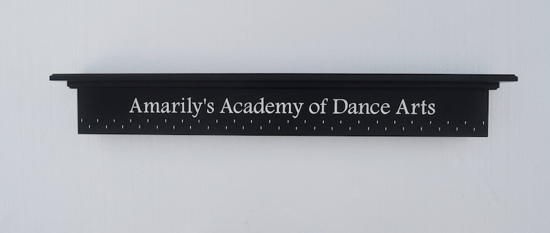 May include: A black shelf with the words "Amarily's Academy of Dance Arts" in white lettering. The shelf has a row of small holes along the bottom.