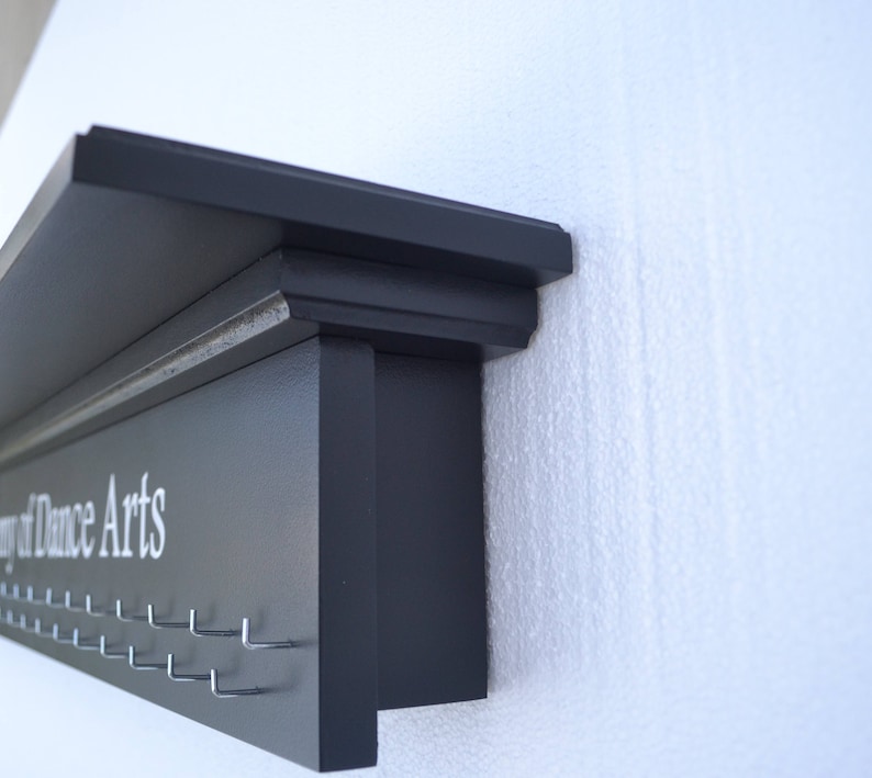 May include: A black wooden shelf with a white inscription that reads "Academy of Dance Arts". The shelf has a decorative molding and multiple hooks for hanging items.