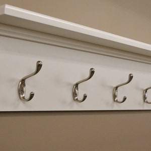 Puede incluir: Estante de madera blanca con cinco ganchos plateados. El estante tiene una moldura decorativa alrededor del borde.