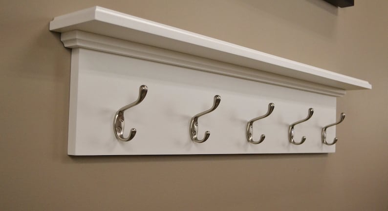 Puede incluir: Estante de madera blanca con cinco ganchos plateados. El estante tiene una moldura decorativa alrededor del borde.