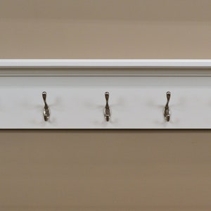 Puede incluir: Estante blanco con cinco ganchos plateados. El estante tiene una moldura decorativa alrededor de la parte superior e inferior.