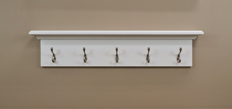 Puede incluir: Estante blanco con cinco ganchos plateados. El estante tiene una moldura decorativa alrededor de la parte superior e inferior.