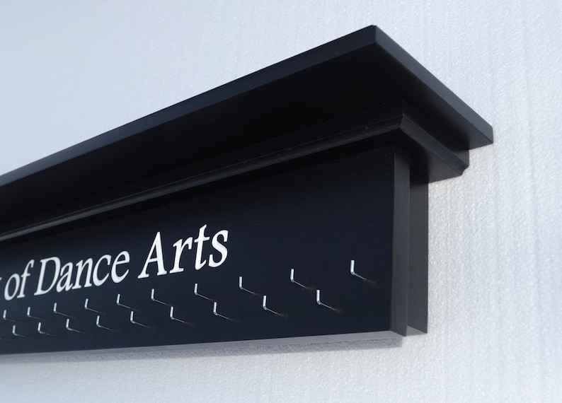 May include: Black wooden wall-mounted medal display with the text "School of Dance Arts" and multiple hooks for hanging medals.