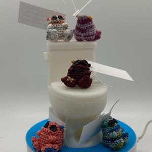 May include: Five crocheted plush toys in various colours, including pink, blue, green, purple, and brown, are sitting on a white toilet and a blue base. The toys are wearing sunglasses and have a whimsical, cartoonish appearance.