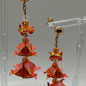 Może przedstawiać: Para kolczyków wiszących z trzema origami-stylowymi kwiatami z papieru w odcieniach pomarańczowego i różowego. Każdy kwiat jest ozdobiony małą złotą koralikiem i przezroczystym koralikiem kryształowym. Kolczyki są zawieszone na złotych haczykach.