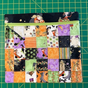 May include: A rectangular zippered pouch featuring a patchwork design of various Halloween-themed fabrics in orange, purple, black, and green. The top section is a solid black fabric with illustrations of witches, while the zipper is lime green.