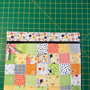 May include: A rectangular fabric pouch featuring a patchwork design of various yellow, green, orange, and white squares with bee, floral, and geometric patterns. The top section has a floral border and a black zipper with a matching fabric pull.