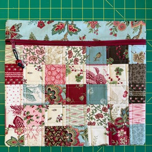 May include: A rectangular fabric pouch featuring a patchwork design of various floral and botanical patterns in shades of red, pink, cream, and sage green. The top section is a solid light blue floral fabric with a dark red zipper and a small bead pull.