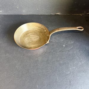 May include: A round copper skillet with a metallic interior finish and a long, textured handle. The pan features a polished copper exterior and a riveted handle attachment, showcasing a classic design with a circular opening at the end of the handle.
