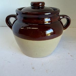 May include: A two-handled ceramic bean pot with a glossy dark brown glazed upper section and a matte beige lower section. The pot features a matching removable lid with a central knob handle and two curved side handles for easy lifting.