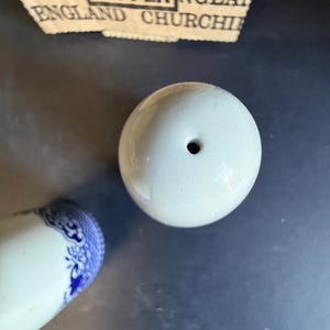 Peut inclure: Un sali&egrave;re en c&eacute;ramique blanche avec un seul trou sur le dessus. La base d'une sali&egrave;re assortie avec des d&eacute;tails floraux bleus est visible. Le haut d'une &eacute;tiquette en papier avec le texte "SALT & PEPPER ENGLAND CHURCH" est en arri&egrave;re-plan.