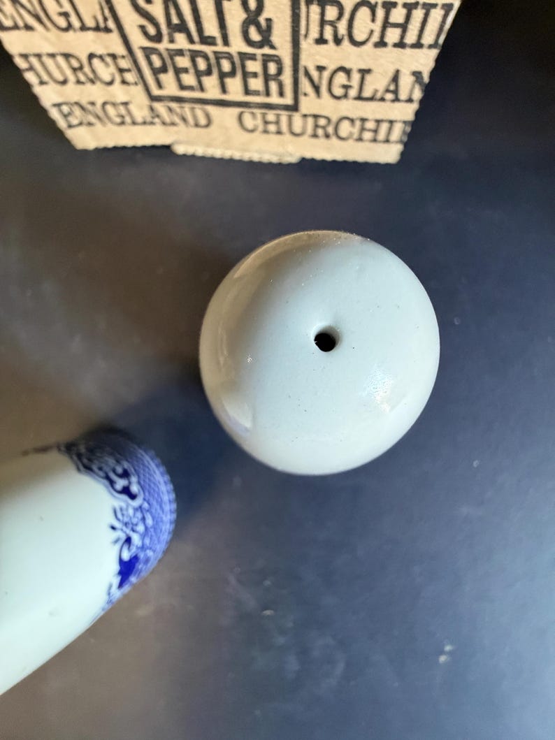 Peut inclure: Un sali&egrave;re en c&eacute;ramique blanche avec un seul trou sur le dessus. La base d'une sali&egrave;re assortie avec des d&eacute;tails floraux bleus est visible. Le haut d'une &eacute;tiquette en papier avec le texte "SALT & PEPPER ENGLAND CHURCH" est en arri&egrave;re-plan.