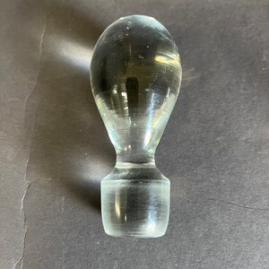 May include: A clear glass bottle stopper with a rounded top and a cylindrical base.