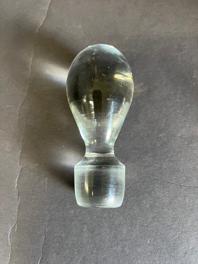 May include: A clear glass bottle stopper with a rounded top and a cylindrical base.