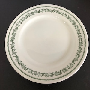 May include: A white ceramic dinner plate with a green floral border. The border features a repeating pattern of small green flowers and leaves.
