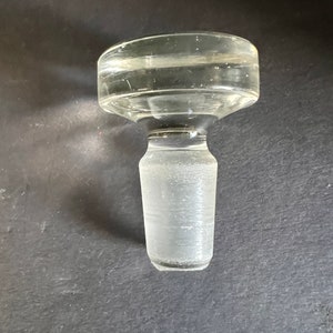 May include: A clear glass bottle stopper with a round top and a cylindrical base. The top has two ridges.