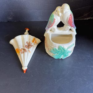 May include: Two ceramic bird figurines perched above a small planter, with colorful wings and a cream-colored body. Beside it, a cream-colored wall pocket with a leaf and autumn leaf design.