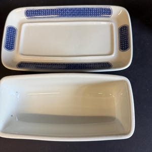 Peut inclure: Un beurrier rectangulaire blanc avec un couvercle, orn&eacute; de motifs bleus sur les bords. Le plat est en c&eacute;ramique et pr&eacute;sente un design classique. La bo&icirc;te au-dessus indique "Covered Butter Churchill of England".