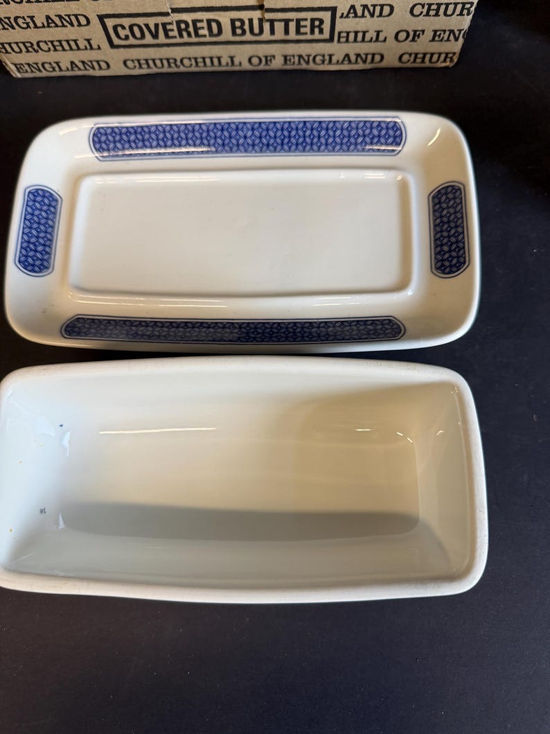 Peut inclure: Un beurrier rectangulaire blanc avec un couvercle, orn&eacute; de motifs bleus sur les bords. Le plat est en c&eacute;ramique et pr&eacute;sente un design classique. La bo&icirc;te au-dessus indique "Covered Butter Churchill of England".