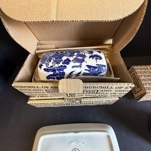 Peut inclure: Un beurrier en c&eacute;ramique bleu et blanc avec un couvercle, emball&eacute; dans une bo&icirc;te en carton. Le plat pr&eacute;sente un motif de paysage d&eacute;taill&eacute;. La bo&icirc;te est &eacute;tiquet&eacute;e "Churchill of England".