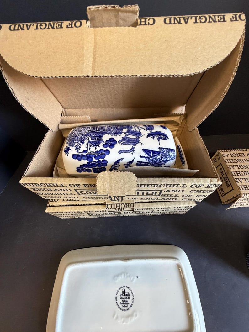 Peut inclure: Un beurrier en c&eacute;ramique bleu et blanc avec un couvercle, emball&eacute; dans une bo&icirc;te en carton. Le plat pr&eacute;sente un motif de paysage d&eacute;taill&eacute;. La bo&icirc;te est &eacute;tiquet&eacute;e "Churchill of England".