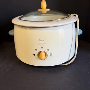 May include: A white Black & Decker slow cooker with a glass lid and yellow accents. The appliance has a control dial and a power cord. The slow cooker has blue handles and the brand name is visible on the front.
