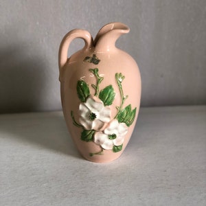 May include: A small, pink ceramic pitcher with a floral design. The pitcher has a white glaze and features white flowers with green leaves.