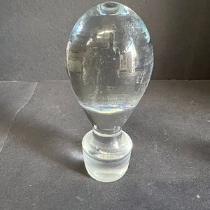 May include: A clear glass bottle stopper with a round, bulbous top and a cylindrical base.