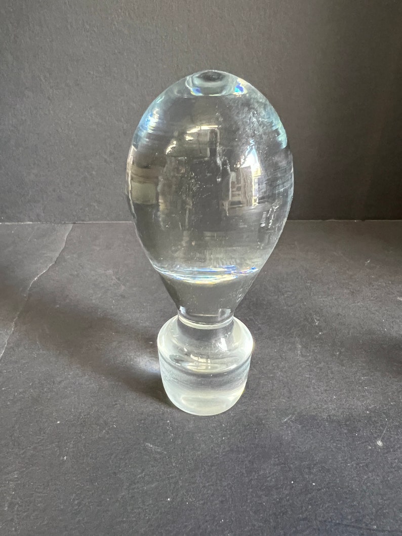 May include: A clear glass bottle stopper with a round, bulbous top and a cylindrical base.